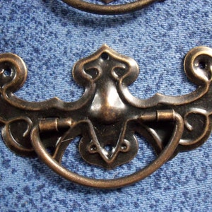 Pode incluir: Uma puxador de armário decorativo de cor bronze com um design ornamentado. O puxador tem uma alça de laço e está preso a uma placa de metal.
