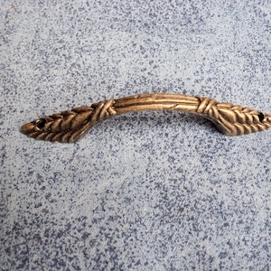 May include: A gold-toned metal cabinet pull with a decorative, woven design. The pull has a curved shape and a slightly pointed end on each side.