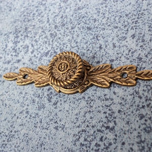 May include: A gold-toned metal drawer pull with a decorative floral design. The pull has a round center piece with a rope-like border and a floral pattern. The pull is attached to a long, rectangular base with a leaf design.