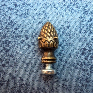 May include: A gold-colored metal finial with a pine cone design. The finial has a silver-colored base with a threaded screw.