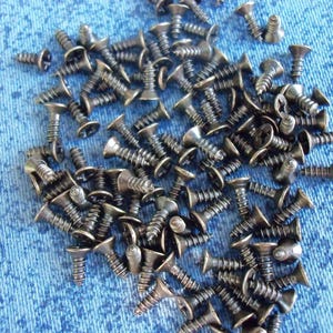 Può includere: Un mucchio di piccole viti metalliche scure con teste piatte e gambi filettati. Le viti sono sparse su una superficie blu strutturata, probabilmente in tessuto. Si tratta di articoli di ferramenta.