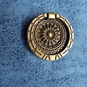 Peut inclure: Un bouton de tiroir rond en laiton antique avec un motif de soleil détaillé au centre. Le bouton a une bordure décorative avec des motifs géométriques et circulaires. Le fond est un tissu bleu texturé.
