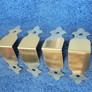 May include: Four gold-colored metal corner brackets with decorative edges. The brackets are designed to be used for furniture or other projects.
