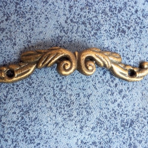 May include: A gold-toned metal decorative accent with a scroll design. The accent has two holes on each side.