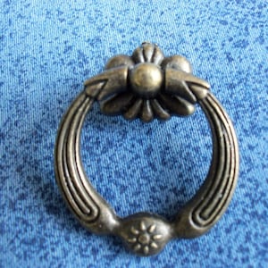 May include: A bronze-colored ring pull with a decorative floral design. The ring pull has a smooth, rounded surface and a slightly raised edge.