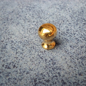 May include: A gold-colored metal knob with a round top and a slightly flared base. The knob is sitting on a blue textured surface.