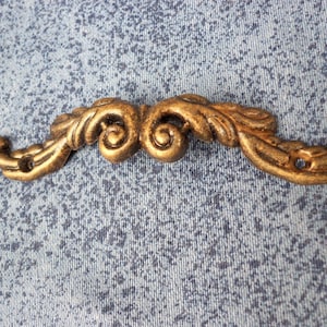 Puede incluir: Una pieza decorativa de metal de color dorado con un diseño de espiral y hojas. La pieza tiene forma de bigote estilizado.