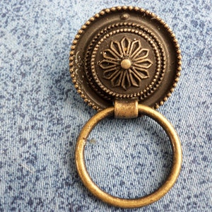 Peut inclure: Une poignée de porte décorative en bronze avec un motif circulaire représentant une fleur stylisée et une poignée en anneau.
