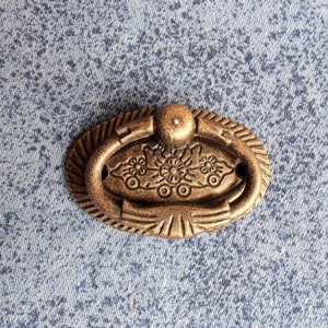 Peut inclure: Une poignée de tiroir ovale en ton or, avec un motif floral décoratif et une poignée en anneau.