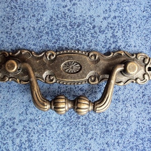 Puede incluir: Un tirador de cajón de bronce de estilo antiguo decorativo con detalles ornamentados y un asa curva.