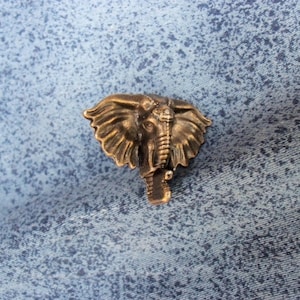 May include: A detailed bronze elephant head pendant. The pendant features a textured surface and intricate details of the elephant's face, including its trunk, tusks, and ears. The pendant is set against a blue background.