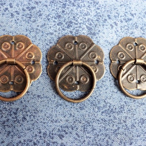 Peut inclure: Trois poignées de tiroir en bronze de style antique avec un motif floral décoratif et une poignée en anneau.