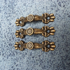 Puede incluir: Tres tiradores de cajón de bronce de estilo antiguo con diseños ornamentados. Cada tirador tiene un pomo hexagonal con un patrón circular en relieve.