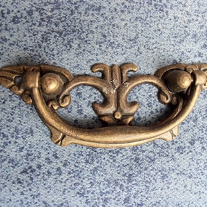 Peut inclure: Une poignée de tiroir en métal doré avec un design décoratif et orné. La poignée a une forme incurvée et une section centrale surélevée avec un motif floral.