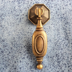 May include: A gold-toned metal cabinet pull with an oval-shaped handle and a decorative top. The pull has a distressed finish.