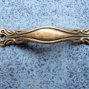 Könnte beinhalten: Ein verzierter, messingfarbener Schubladenknauf im Vintage-Stil mit einem dekorativen, gerollten Design. Der Knauf hat eine glatte, ovale Mitte.
