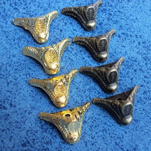 Peut inclure: Un assortiment de pieds de meubles décoratifs en or et bronze. Les pieds ont une forme triangulaire avec des détails ornementaux en forme de feuilles. Ils sont disposés sur une surface bleue texturée.