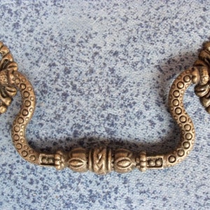 Puede incluir: Una manilla de tiro decorativa, adornada, de metal con un diseño de remolino y textura. La manilla es de color dorado claro y tiene un diseño floral en cada extremo.