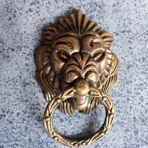 Puede incluir: Un llamador de puerta de bronce con forma de cabeza de león con un asa de anillo. La cabeza de león está detallada con una melena texturizada y una expresión feroz.