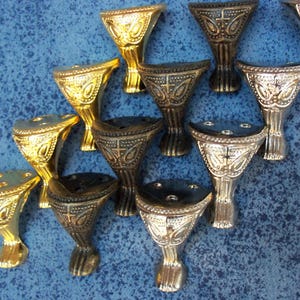 May include: A selection of decorative furniture feet in gold, silver, and bronze finishes. Each foot has an ornate design with a flared top and a claw-like base. The feet are displayed on a blue surface. The feet are likely for a table or chair.