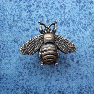 Puede incluir: Adorno metálico con forma de abeja de estilo antiguo. La abeja presenta alas y segmentos corporales detallados, con un aspecto texturizado y envejecido. La abeja está sobre un fondo azul texturizado.