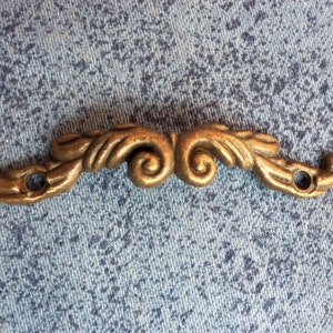 Puede incluir: Un tirador de cajón de metal dorado con un diseño decorativo y ornamentado. El tirador tiene dos agujeros para el montaje.