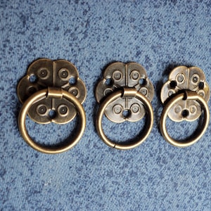 May include: Three antique-style brass drawer pulls with a decorative floral design and a ring handle. The pulls are arranged in a row on a blue fabric background.