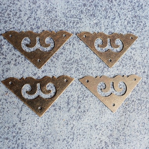 May include: Four antique bronze-colored corner brackets with decorative scrollwork. Each bracket is triangular with three screw holes and a stylized design. The brackets are arranged on a textured blue-gray surface.