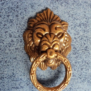 May include: A gold lion's head door knocker with a ring handle. The knocker is detailed with a textured mane and a fierce expression.