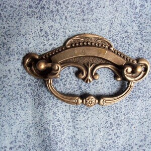 May include: A vintage-style, ornate, brass drawer pull with a decorative scroll design and a blank rectangular plate.