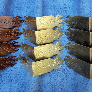 May include: An assortment of decorative metal corner brackets in various finishes, including bronze and gold. Each bracket features an ornate floral design and is designed to protect and embellish corners. The brackets are arranged on a blue surface.