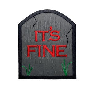 May include: A gray embroidered patch shaped like a tombstone with the text "IT'S FINE" in red lettering and green grass on the bottom.