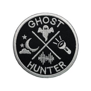 May include: Black and white embroidered patch with the text "Ghost Hunter" in a circle. The patch features a ghost, a crescent moon, a flashlight, and a sound wave.