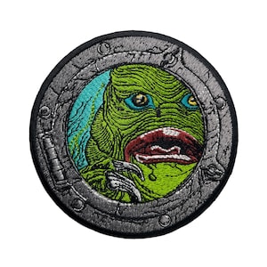 May include: A circular embroidered patch with a black border and a silver-grey interior. The patch features a green, blue, and red creature with a large mouth and sharp teeth, peering through a round porthole.