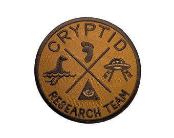 Puede incluir: Un parche bordado marrón con las palabras "Cryptid Research Team" en un círculo alrededor de una cruz con una huella, un monstruo del Lago Ness, un OVNI y un ojo que todo lo ve.