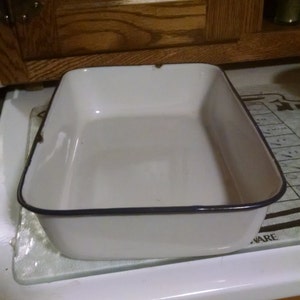 May include: A rectangular white enamel baking dish with a dark blue rim. The dish is empty and sits on a glass cutting board. The kitchen cabinet is visible in the background.