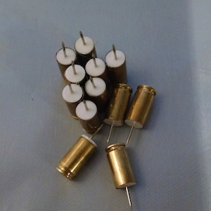 May include: A group of 10 brass push pins with white plastic heads and silver metal points. The push pins are scattered on a light blue fabric surface.