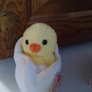 May include: A plush yellow chick with black eyes and an orange beak sits inside a white plush egg. The chick is a stuffed animal. The egg has a red and white tag attached to it. The background is a wooden surface.