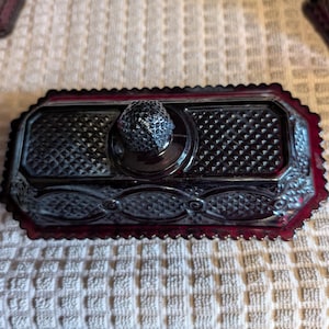 May include: A rectangular dark red glass butter dish with a lid featuring a faceted knob handle. The glass surface displays a textured diamond pattern across the top and sides, finished with a scalloped edge along the base of the lid.