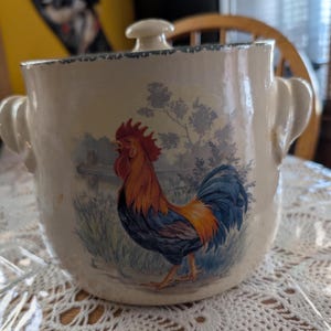 May include: Cream-colored ceramic container with a lid and handles. The container features a vibrant illustration of a rooster with red, orange, and blue plumage, set against a pastoral scene. The rim and lid are trimmed with a blue floral pattern.