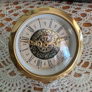 May include: A round table clock with a polished gold-tone metal frame and a glass cover. The clock face features a white outer ring with black Roman numerals, surrounding a central decorative gold and black filigree pattern with ornate gold hands.