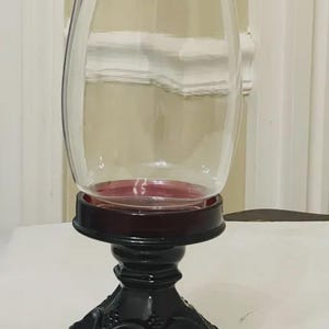 May include: A clear glass hurricane shade sits atop a dark red, pedestal-style candle holder. The base features a textured, pressed glass pattern with a wide, circular foot. The candle holder has a deep red rim where the glass shade rests.
