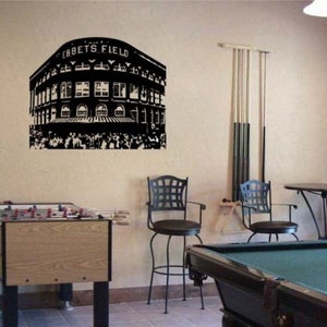 May include: Black and white silhouette of Ebbets Field, a historic baseball stadium, on a beige wall. The image is a wall decal. There is a pool table, two bar stools, and a foosball table in the room.