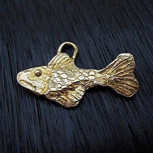 May include: A gold-colored fish pendant with textured scales and a loop for a chain. The pendant is a detailed, three-dimensional design, perfect for jewelry. The fish is lying on a dark, textured surface.