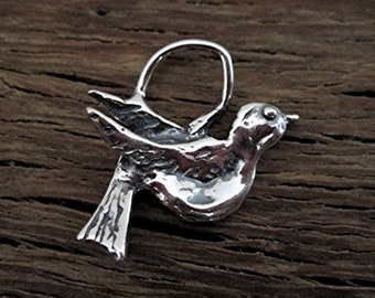 May include: A silver-colored bird pendant with a textured surface. The bird is depicted in flight, with its wings spread and a loop at the top for hanging. The pendant is set against a dark wood grain background.