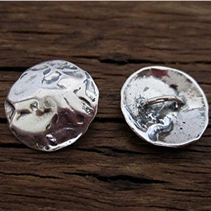 May include: Two silver-colored, irregularly shaped buttons. One button is face up, showing a textured surface. The other button is face down, revealing a loop for attachment. The buttons are on a dark wood surface.