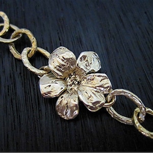 May include: A gold-colored necklace with a flower pendant. The flower is the focal point, with textured petals and a detailed center. The chain links are also gold and have a hammered texture.