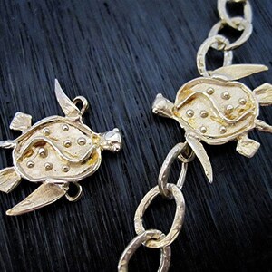 May include: Close-up of two gold-colored turtle pendants, one attached to a chain. The turtles have detailed shells with small, raised dots and flipper-like appendages. The chain links are oval-shaped.