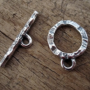 Könnte beinhalten: Silberner Toggle-Verschluss mit strukturierter Oberfläche. Der Verschluss hat eine Stange mit einer kleinen Schlaufe an einem Ende und einen runden Ring mit einer Schlaufe am anderen Ende.
