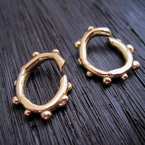 May include: Two gold-colored, open-hoop earrings. Each earring has a C-shape with a series of small, round, raised details along the outer edge. The earrings are displayed on a dark, textured surface.